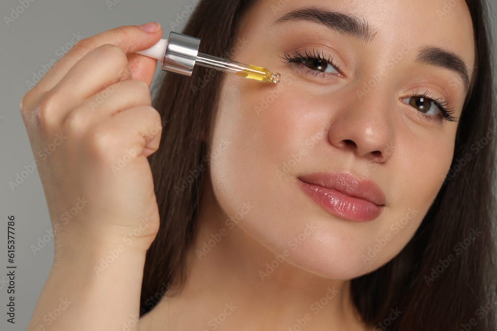 Canvas Prints Beautiful young woman applying essential oil onto face on grey background, closeup