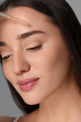 Beautiful young woman applying essential oil onto face on grey background, closeup
