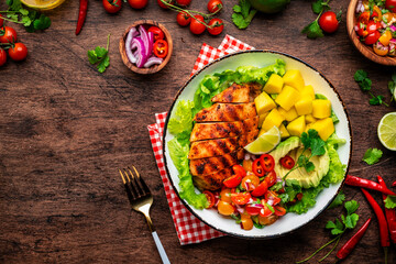 Grilled chicken with side dish salad from avocado, mango, tomato salsa, cilantro, lime and lettuce, rustic wooden table background, top view