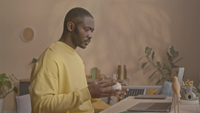 Side view waist up shot of young African American man bouncing baseball back and forth in hands while working on laptop at desk in home office