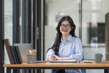 Attractive young Asian businesswoman or female office employee wearing eyeglasses sitting at her office desk, smiling and looking at the camera.