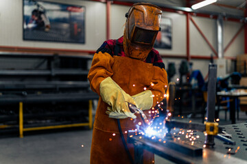 A woman in protective equipment uses tools and machines in the workshop, sparks fly and illuminate...