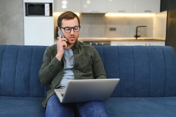 Cheerful busy young caucasian male manager sits on sofa with computer, calls by phone, talks with client, looks at free space in white room interior. Business, work remotely at home, ad and offer.