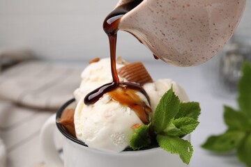 Pouring caramel sauce onto ice cream with candies and mint leaves, closeup
