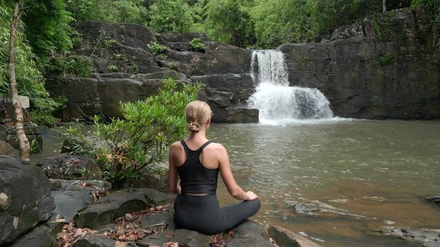 Beautiful young woman practice meditation at a waterfall in the rainforest in Thailand.