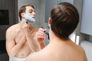Handsome young man is shaving his face and looking at the mirror.