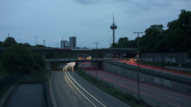 Traffic Flow and Cologne Skyline. Witness bustling cars with light strokes on a highway, a prominent TV tower, moving clouds, and Cologne's panoramic skyline in the evening. Energy and dynamism. 002