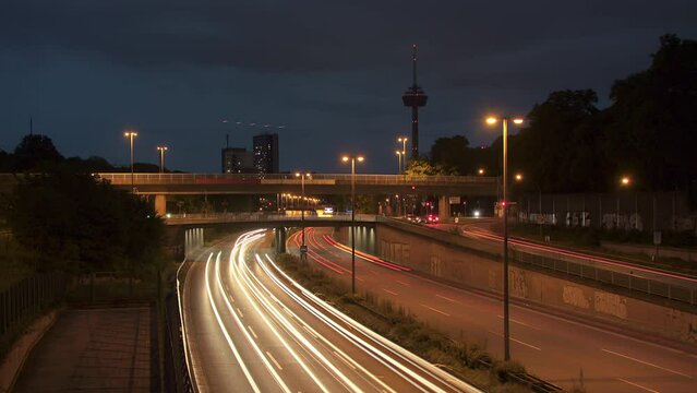 Traffic Flow and Cologne Skyline. Witness bustling cars with light strokes on a highway, a prominent TV tower, moving clouds, and Cologne's panoramic skyline in the evening. Energy and dynamism. 003