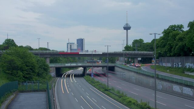 Traffic Flow and Cologne Skyline. Witness bustling cars with light strokes on a highway, a prominent TV tower, moving clouds, and Cologne's panoramic skyline in the evening. Energy and dynamism. Z001