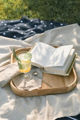 Lemonade garnished with lemon and rosemary twig, open book on wooden tray at a picnic in nature.