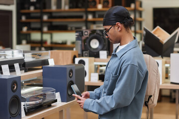 Side view portrait of young black man looking at vinyl record player in music store and taking picture of price tag with smartphone