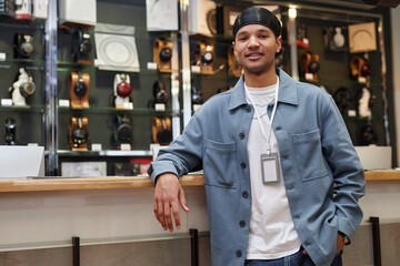 Waist up portrait of smiling young man working in music store and standing by counter looking at...
