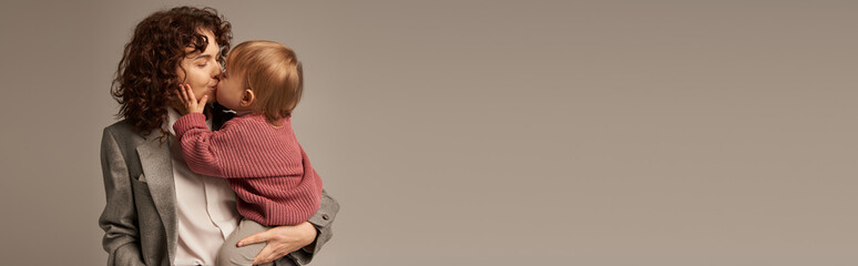 work life balance concept, curly woman in suit kissing daughter on grey background, loving mother,...