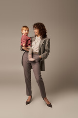 quality time, work life balance concept, smiling woman in suit holding daughter and standing with hand in pocket on grey background, career and family, loving motherhood, full length