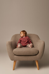 cute baby girl, toddler in casual attire sitting on comfortable armchair on grey background in studio, emotion, confused, innocence, little child, toddler fashion, stylish outfit, sweater