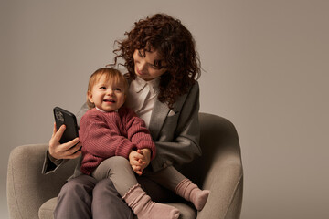 modern working mother, multitasking, using smartphone, balancing work and life concept, happy businesswoman in suit sitting with toddler child on armchair, grey background, parent and child