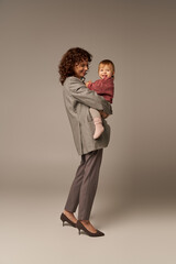 quality family time, parenting and career, joyful businesswoman holding in arms her toddler daughter on grey background, work life harmony concept, working parent, loving motherhood, full length