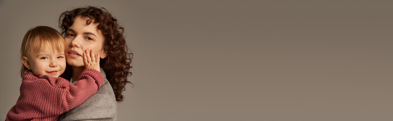 working mother, parenting and career, curly businesswoman embracing toddler child on grey background, work life harmony concept, loving motherhood, quality family time, banner