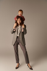 kid on shoulders, loving motherhood, parenting and career, curly businesswoman and happy toddler daughter on grey background, work life harmony concept, loving motherhood, quality family time