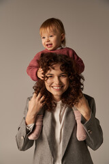 loving motherhood, parenting and career, curly businesswoman and happy toddler daughter on grey background, kid on shoulders, work life harmony concept, loving motherhood, quality family time