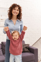 loving motherhood, balancing work and life, family bonding, happy and curly mother lifting toddler daughter, togetherness, cozy living room, quality time, denim jeans, engaging with child