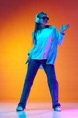 Young woman in casual clothes and sunglasses listening to music in headphones and dancing against gradient orange studio background in neon light