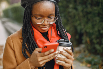 Young black woman in a coat drinking coffee outside and using a phone
