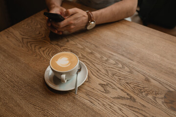 Fototapeta na wymiar Cup of coffee on wooden table next to a man using cell phone. Coffee break in a city cafe.