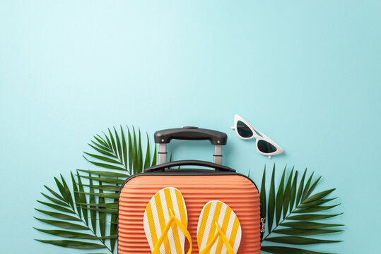 Vacation mode on! Overhead shot of an orange suitcase, beach accessories, sunglasses, flip-flops, and natural palm leaves on a pastel blue backdrop, with space for text or advertising