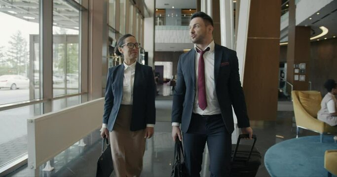 Man and woman in formal wear are walking along hotel lobby with baggage smiling talking laughing enjoying business trip. People and travelling concept.