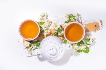 Organic herbal health drink. Jasmine flowers green tea, in white mugs, with teapot, on white table, with blooming jasmine flowers 