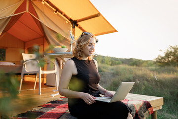 Happy Woman freelancer using a laptop on a cozy glamping tent in a sunny day. Luxury camping tent for outdoor summer holiday and vacation. Lifestyle concept