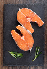 Raw portioned salmon steaks and rosemary sprigs on a mica serving board over a wooden background.
