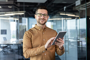 Obraz na płótnie Canvas Portrait of a young male designer standing in the office and using a tablet. He looks at the camera with a smile