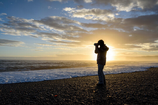 Silhouette of a photographer standing on the beach taking a picture of the North Atlantic Ocean view at sunset; Iceland