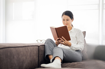 Senior asian woman Reading Book Sitting On Couch At Home. Retired female Enjoying Reading New Novel Or Business Literature On Weekend. Retirement Leisure Concept