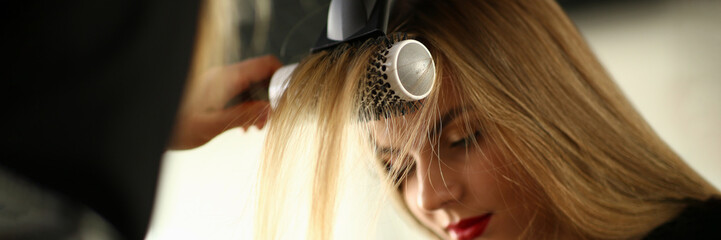 Drying blond hair with a hairdryer and a round brush in barbershop. Master doing hair styling to a...
