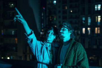 Young couple of lovers stands on city roof in neon lighting against background of urban landscape with high buildings at night.