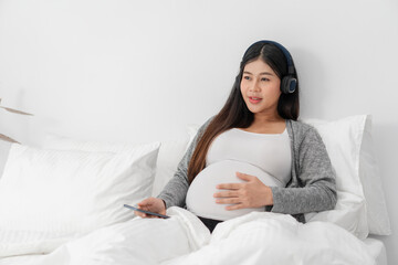 Obraz na płótnie Canvas Asian happy pregnant woman is sitting on bed and and touching her belly and wear a headphone to listen the music. pregnancy, motherhood, people and expectation concept