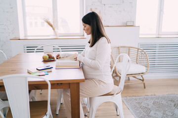Young expectant woman reading book at table n kitchen at home