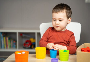 A beautiful little boy of two or three years old sits at a table in a children's room and plays with toys. Multicolored buckets. Pyramid for children. Cute toddler