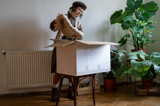 Hipster girl stacking old clothes into cardboard box at home, sorting unwanted items to recycle. Clothing waste and recycling, conscious living concept