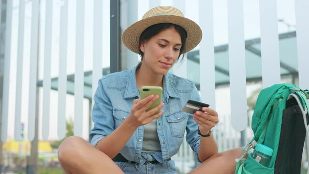 Tourist girl in hat holding credit card and look at cellphone. Bus station background. Young traveller woman at public transport stop trying buy tickets. Female customer buy tickets online.
