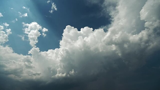 Time lapse of blue sky with billowing storm clouds that have white tops and gray bottoms.  Some sun rays occur in the top left corner.