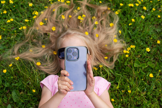 Little girl takes a photos or shoots a video on the smartphone lying on the grass with yellow flowers.