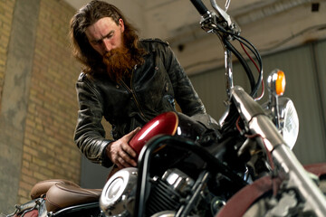 Plakat Creative authentic motorcycle workshop Garage of red bearded biker mechanic wiping beautiful motorcycle with rag