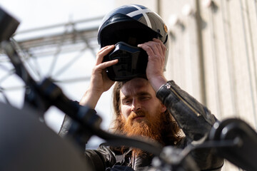 brutal red bearded biker sitting on a black motorcycle after a race takes off his helmet lust for...