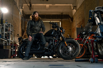 Fototapeta na wymiar Creative authentic motorcycle workshop garage serious redhead bearded biker mechanic sitting on motorcycle