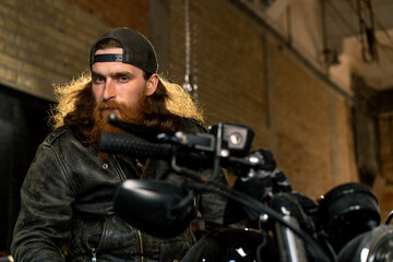 Obraz na płótnie Canvas Creative authentic motorcycle workshop garage portrait serious redhead bearded biker mechanic sitting on motorcycle