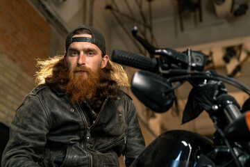 Creative authentic motorcycle workshop garage portrait serious redhead bearded biker mechanic sitting on motorcycle